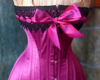 May include: A magenta satin corset with black lace trim and a large magenta satin bow. The corset has a busk closure and is designed to cinch the waist.