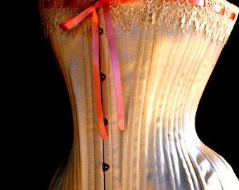 May include: A white satin corset with a pink ribbon and lace trim. The corset has a boned structure and a hook and eye closure in the back.