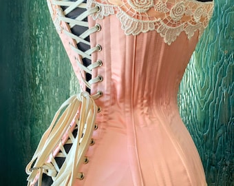 May include: A peach-colored corset with ivory lacing and trim. The corset features a delicate ivory lace trim along the top edge, with a floral pattern. The corset is displayed on a black mannequin, showcasing its shape and design.