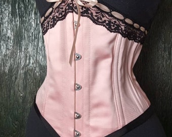 May include: A pink satin corset with black lace trim and a satin ribbon bow. The corset has metal hooks and eyes for closure.
