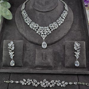 Puede incluir: Un deslumbrante conjunto de joyas que incluye collar, pendientes y pulsera. El conjunto está adornado con piedras claras y brillantes dispuestas en un patrón floral. El collar tiene un colgante en forma de lágrima, y los pendientes cuelgan elegantemente. La pulsera refleja el diseño del collar.