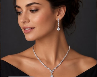 May include: A silver-toned necklace and matching drop earrings set featuring clear, faceted stones. The necklace has a central pear-shaped stone pendant, while the earrings consist of a vertical arrangement of small round stones and a pear-shaped drop.
