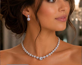 May include: A silver-toned necklace featuring a continuous row of round, clear, faceted stones that increase in size toward the center, paired with matching drop earrings consisting of a single round stone suspended from a smaller stone.