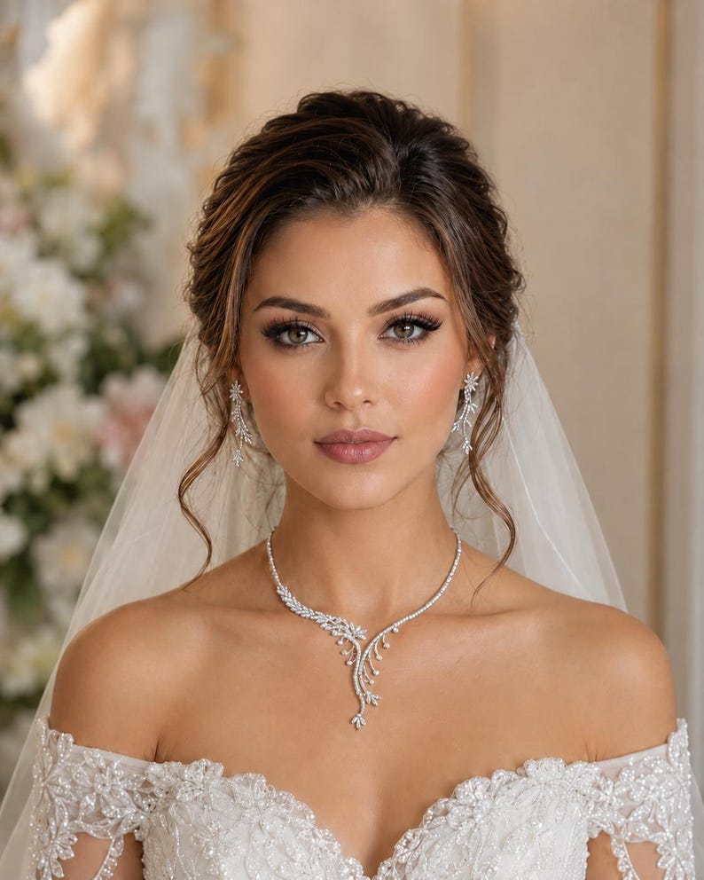 May include: A silver-toned necklace and matching drop earrings featuring a delicate leaf-like design encrusted with small clear stones. The jewelry set is worn with an off-the-shoulder white lace dress and a sheer white veil, creating a cohesive look.