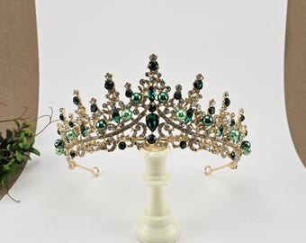 May include: A gold crown with green and clear rhinestones and pearls. The crown is on a white stand.