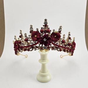May include: A gold crown with red fabric flowers and rhinestones. The crown is on a white stand.