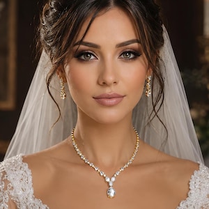 May include: A gold-toned necklace featuring a central pear-shaped clear stone surrounded by smaller round clear stones, paired with matching drop earrings. The jewelry set is worn with a white lace garment and a sheer white veil.