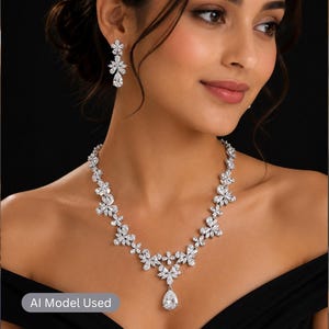 May include: A silver-toned necklace and matching drop earrings set featuring clear, faceted stones arranged in floral-inspired clusters. The necklace includes a central pear-shaped stone pendant, and the earrings mirror the floral design.