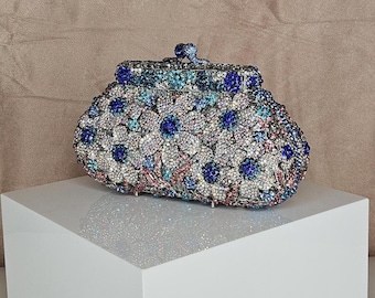 May include: A sparkling, floral-patterned clutch in shades of blue, silver, and pink. The evening bag is covered in small, glittering embellishments, with a clasp closure. The clutch is displayed on a white, geometric pedestal.