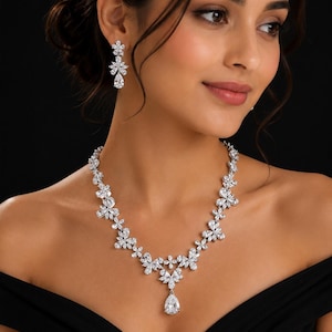 May include: A silver-toned necklace and matching drop earrings set featuring clear, faceted stones arranged in floral patterns. The necklace consists of multiple connected flower motifs, with a single pear-shaped stone pendant at the center.