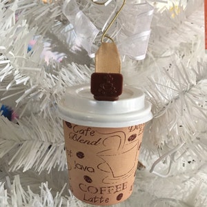 May include: A coffee cup ornament with a white lid and brown paper cup. The cup has coffee-related text and graphics. A brown charm with a coffee cup design and a gold wire hanger with white ribbon complete the ornament.