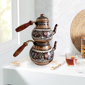 May include: A traditional Turkish tea set with a copper teapot decorated with colorful floral patterns. The teapot has two sections and wooden handles. The set is displayed on a white countertop.