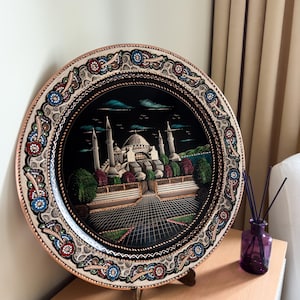 Può includere: Un piatto rotondo in rame con sfondo nero e una pittura dettagliata di una moschea con minareti. Il piatto ha un bordo decorativo con motivi floreali in rosso, blu e bianco.