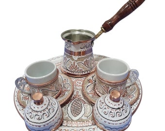 May include: A traditional Turkish coffee set with a copper pot, two cups, and saucers, and two lids. The set features intricate floral designs in white and gold on a white background.
