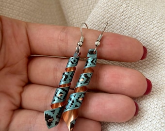 May include: A pair of copper and turquoise earrings with a geometric pattern. The earrings are made of copper wire that has been coiled and then decorated with a turquoise and black geometric pattern. The earrings are suspended from silver hooks.