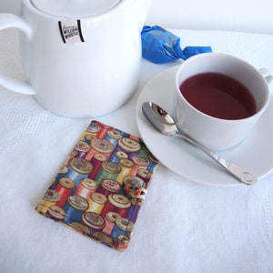 May include: A white teapot with a black label that says "William Whistle" on it. The teapot is sitting on a white tablecloth next to a white teacup with a red liquid inside. There is a blue and silver wrapped sweet next to the teapot. A colourful fabric tea cosy with a button is in the foreground.