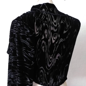 May include: A black velvet bolero jacket with a distinctive swirling pattern in a lighter shade of black. The jacket features long sleeves and a high collar, providing a sophisticated and elegant appearance. The fabric looks soft and drapes well.