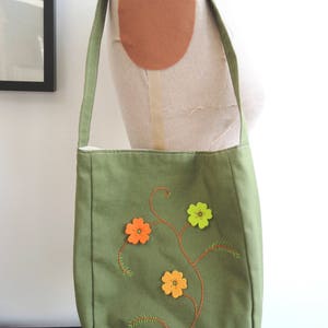 May include: A green canvas tote bag with a floral design embroidered on the front. The flowers are orange, yellow, and green.