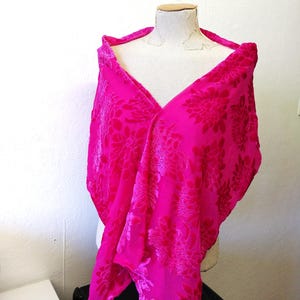 May include: A bright fuchsia shawl with a floral pattern, displayed on a mannequin. The fabric has a velvety texture, and the floral design is a darker shade of fuchsia. A striking accessory.
