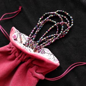 May include: A deep pink velvet drawstring bag with a patterned lining, holding strands of iridescent beads. The bag's drawstring is also deep pink. The beads are a mix of colours, reflecting light. The background is black.