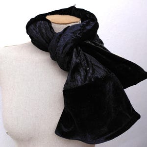 May include: A black velvet scarf displayed on a mannequin. The scarf is made of a soft, plush material and has a deep, dark colour. The scarf is knotted at the neck and has a wide, flared end. The background is a plain white wall.