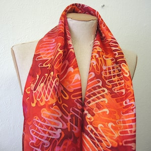May include: A red, orange and pink silk scarf with a swirling pattern. The scarf is draped over a mannequin's neck.