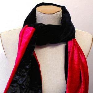 May include: A black velvet scarf with a bright red lining is draped over a mannequin. The scarf is twisted at the neck, showing the contrast between the black exterior and the bright red interior. The fabric appears soft and luxurious.