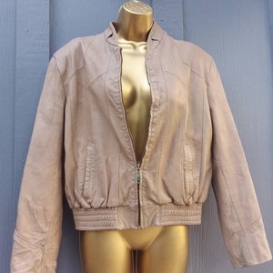 May include: A tan leather jacket with a zip-up front, displayed on a gold mannequin. The jacket has a ribbed hem and cuffs, and a stand-up collar. The jacket is a neutral color and appears to be vintage.