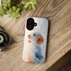 May include: A white phone case with a white and orange dog illustration. The dog is wearing a white and blue scarf. The phone case has a black camera lens.