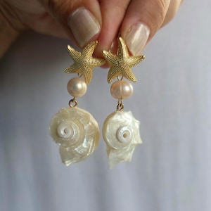 May include: A pair of drop earrings featuring a gold-toned textured starfish at the top, followed by a single round cream-colored pearl, and finished with a dangling white iridescent spiral seashell charm with a smooth, pearlescent finish.