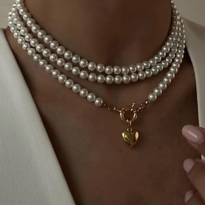 May include: A layered pearl necklace with a gold heart pendant. The necklace features three strands of white pearls and a gold clasp. The heart pendant is a solid gold color.