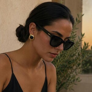 May include: Black square-framed sunglasses with dark lenses and a gold-tone metal detail on the temple. The eyewear is paired with a gold-tone square stud earring featuring a central black inlay and a textured, rope-like border design.
