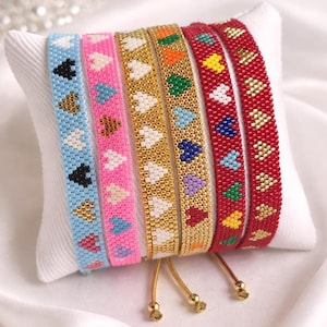 Pode incluir: Seis pulseiras de contas em várias cores, cada uma adornada com padrões de corações. As pulseiras são exibidas em uma almofada branca. As cores incluem azul claro, rosa, dourado e vermelho, com desenhos de corações em cores contrastantes.
