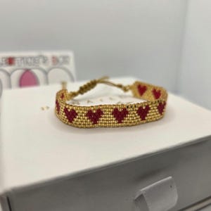 May include: A gold beaded bracelet with a repeating pattern of red hearts. The bracelet is adjustable with a woven cord closure. The bracelet is displayed on a white surface.