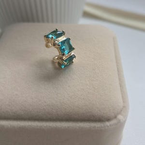 May include: A gold-colored ear cuff featuring three square, light blue gemstones. The ear cuff is displayed on a beige surface, showcasing the jewelry's design and color.