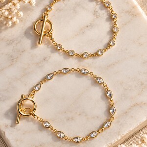 May include: Two gold-toned bracelets featuring a toggle clasp. The top bracelet has round clear crystals set in gold links. The bottom bracelet features teardrop-shaped clear crystals set in gold links. Both pieces have a polished metallic finish.