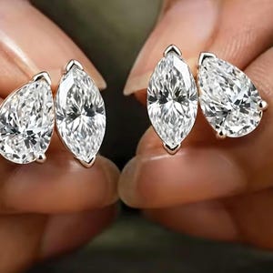 Puede incluir: Un par de pendientes de botón, cada uno con una piedra preciosa transparente en forma de pera y una piedra transparente de talla marquesa engastadas en un marco metálico. Las piedras facetadas reflejan la luz, creando un aspecto brillante.