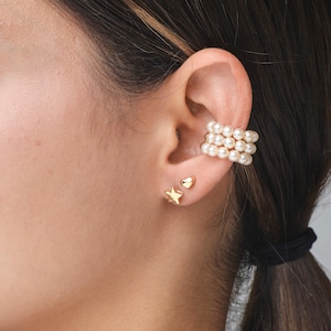 May include: Earrings set featuring a gold star and heart stud, and a pearl ear cuff. The ear cuff has three rows of pearls. The earrings are worn on a person's ear.