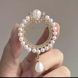 May include: A gold-toned brooch with a circular design, adorned with small white pearls and clear crystals. A large pearl sits at the top, and a teardrop-shaped pearl dangles from the bottom. The brooch is held in a hand.