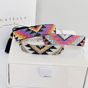 May include: Three beaded bracelets with geometric patterns in various colors. One bracelet features black, yellow, orange, and purple beads. Another has pink, orange, and gray beads. The third bracelet has black, gold, and white beads. The bracelets are displayed on a white box.