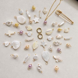 Puede incluir: Una colección de componentes de joyería que incluye dijes de nácar blanco en forma de corazón, estrella y lágrima, cuentas redondas doradas y lavanda, dos aros dorados, un imperdible dorado y una delicada cadena dorada con una cuenta lavanda.