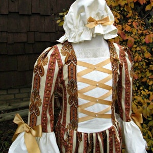 May include: A white dress with a brown and pink paisley print. The dress has a white bodice with a criss-cross ribbon detail and a white lace trim at the cuffs. The dress is worn with a white bonnet with a gold ribbon.