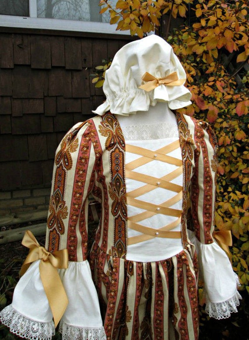 May include: A white dress with a brown and pink paisley print. The dress has a white bodice with a criss-cross ribbon detail and a white lace trim at the cuffs. The dress is worn with a white bonnet with a gold ribbon.