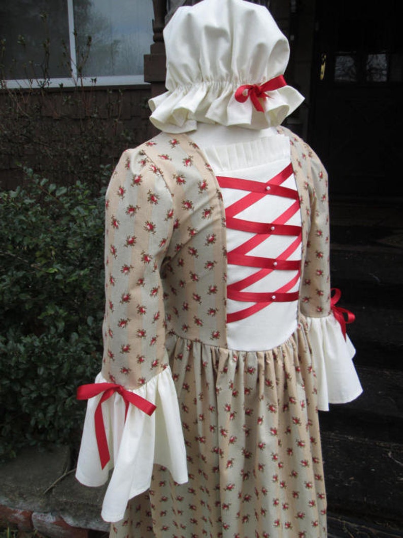 May include: A beige and white dress with a floral print and red ribbon accents. The dress has a white lace collar and a white cap with a red ribbon.