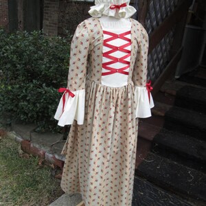 May include: A white dress with a red ribbon criss-cross design on the back. The dress has a floral pattern and a white bonnet with a red ribbon.