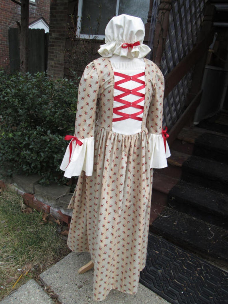 May include: A white dress with a red ribbon criss-cross design on the back. The dress has a floral pattern and a white bonnet with a red ribbon.
