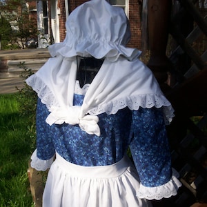 May include: A white bonnet with a ruffled edge and a white lace trimmed apron over a blue floral print dress.