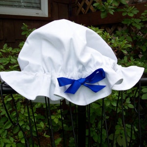 May include: A white bonnet with a ruffled brim and a blue satin bow. The bonnet is made of a soft, lightweight fabric and is likely for a child. The design is reminiscent of historical or vintage styles.