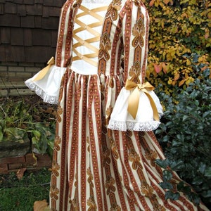 May include: A white and brown striped dress with gold accents and a lace trim. The dress has a fitted bodice with a criss-cross ribbon detail and a full skirt.