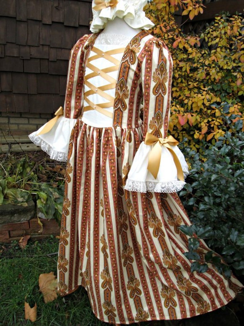 May include: A white and brown striped dress with gold accents and a lace trim. The dress has a fitted bodice with a criss-cross ribbon detail and a full skirt.